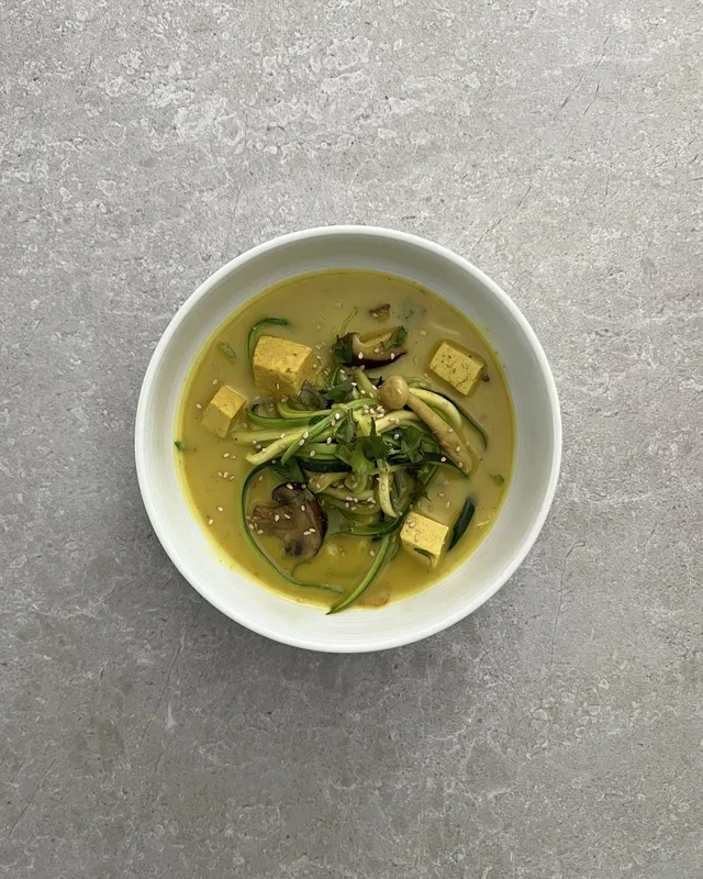Bol blanc amb fideus de carbassó, daus de tofu, bolets, coriandre i sèsam en un brou groc de llet de coco