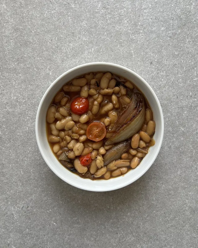 Bol blanco con alubias blancas en su caldo, cuartos de cebolla asada y tomates cherry partidos por la mitad